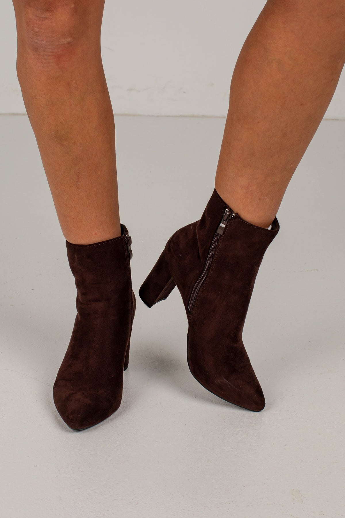 A pair of dark brown suede ankle boots with pointed toes and block heels, shown on a model's feet against a light gray studio background.