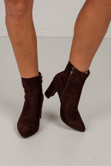 A pair of dark brown suede ankle boots with pointed toes and block heels, shown on a model's feet against a light gray studio background.