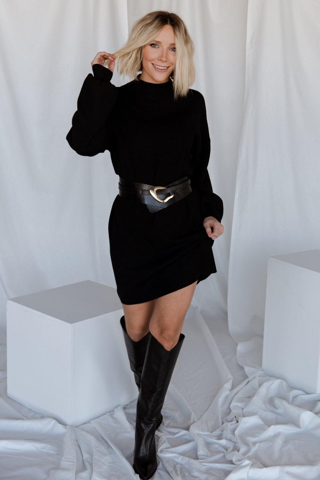 Front view of a short black sweater dress with mock neckline and long bubble sleeves on a model, styled with ankle boots and minimal accessories.