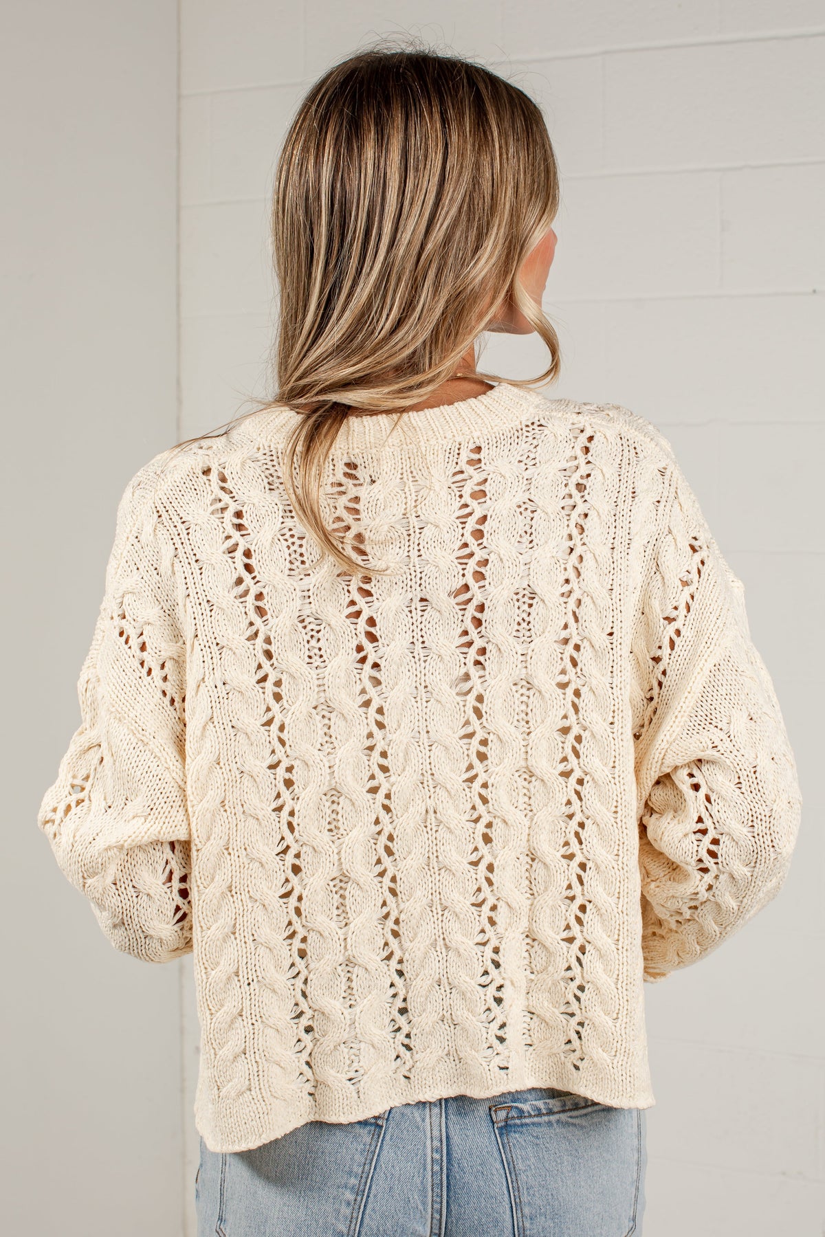 A woman viewed from behind wearing a cream-colored, cable-knit cropped sweater with textured openwork patterns, paired with light blue jeans, against a light gray brick wall.