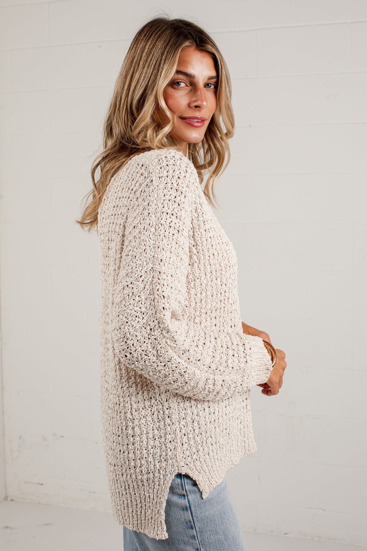 A woman wearing a chunky, cream-colored open-knit sweater and light blue jeans, standing against a white paneled wall.