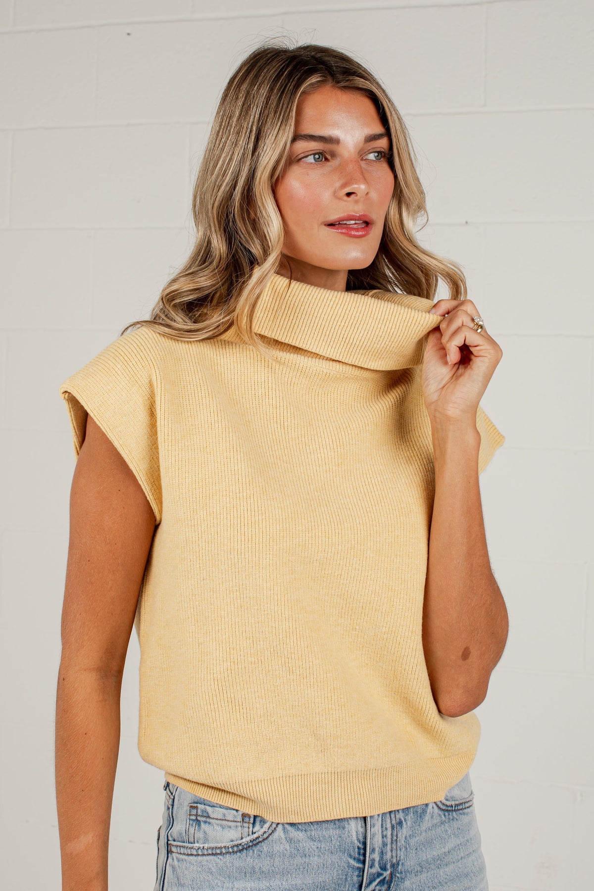 A woman wearing a pale yellow ribbed knit turtleneck sweater with cap sleeves, paired with light blue jeans, standing against a white brick wall.