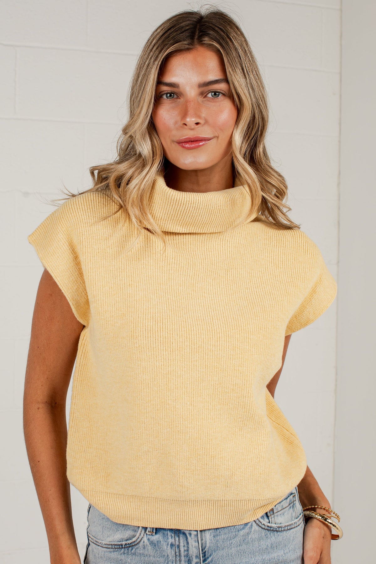 A woman wearing a pale yellow ribbed knit mock neck top with cap sleeves and light blue jeans, standing against a white brick wall.