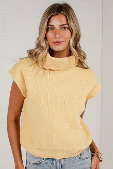 A woman wearing a pale yellow ribbed knit mock neck top with cap sleeves and light blue jeans, standing against a white brick wall.
