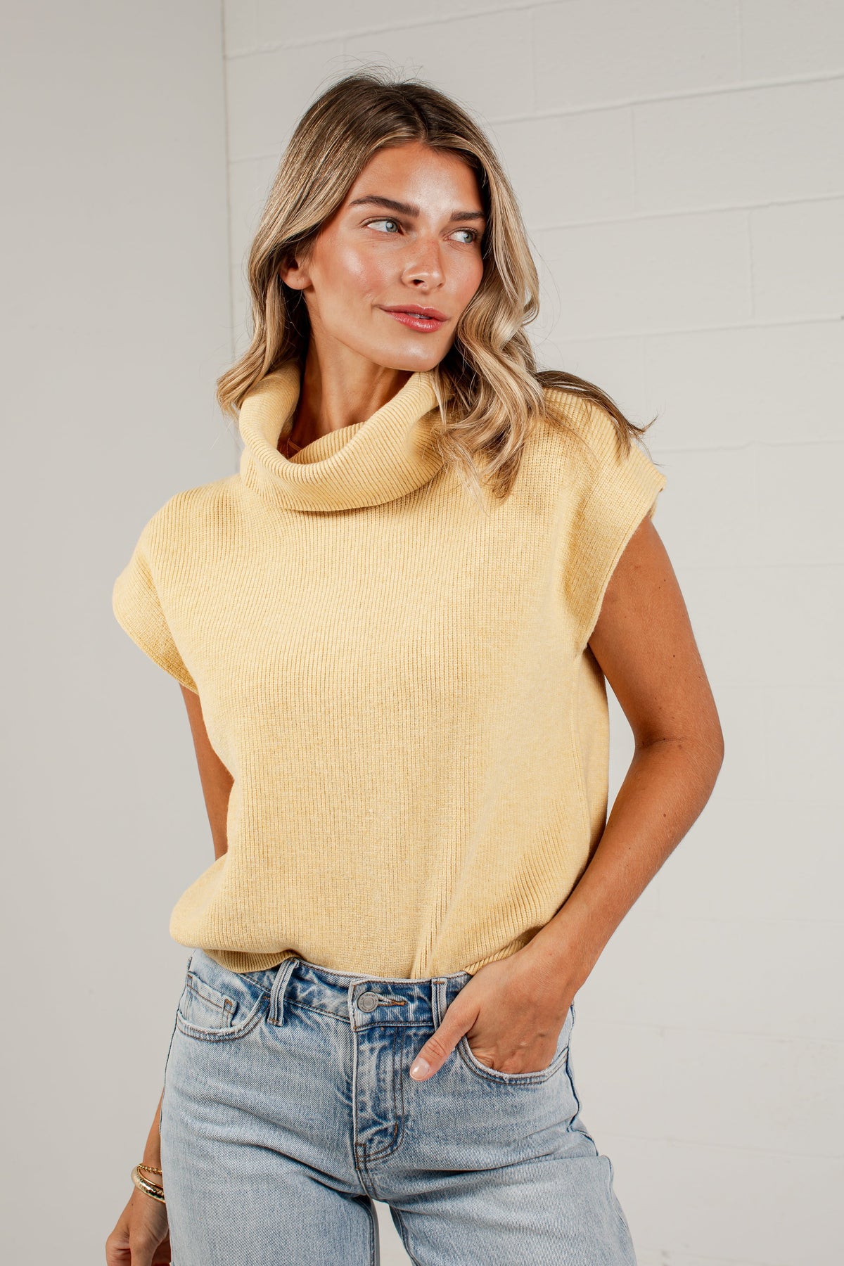 A woman wearing a pale yellow ribbed knit turtleneck sweater with cap sleeves and light-wash blue jeans, standing against a white brick wall.