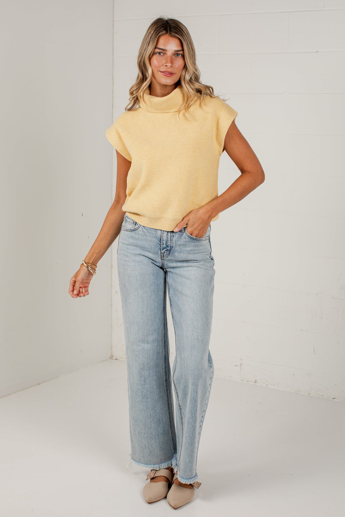 A woman modeling a pale yellow knit mock-neck top with short sleeves and light-wash wide-leg jeans, standing against a white brick wall.